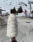 Person in a white fur coat standing on a snowy mountain with ski lifts and mountains in the background.